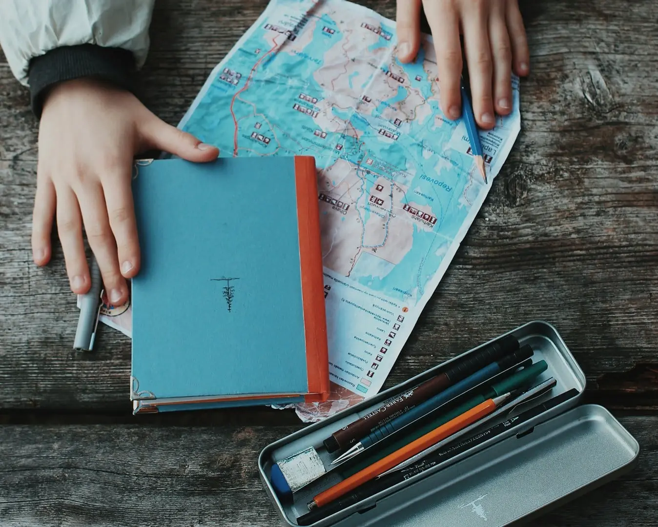 Manos sobre un mapa de papel y un cuaderno de viaje, junto a un estuche de lápices